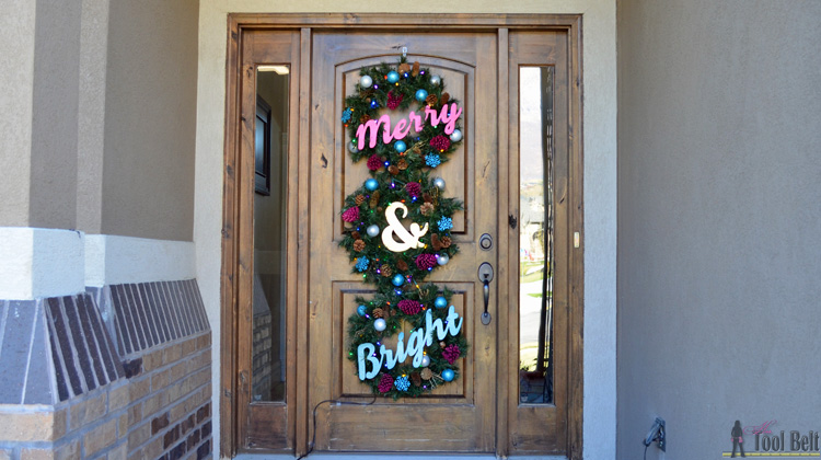 Three evergreen wreaths stacked down a tall wood front door with plywood letters reading Merry and Bright in pink and teal